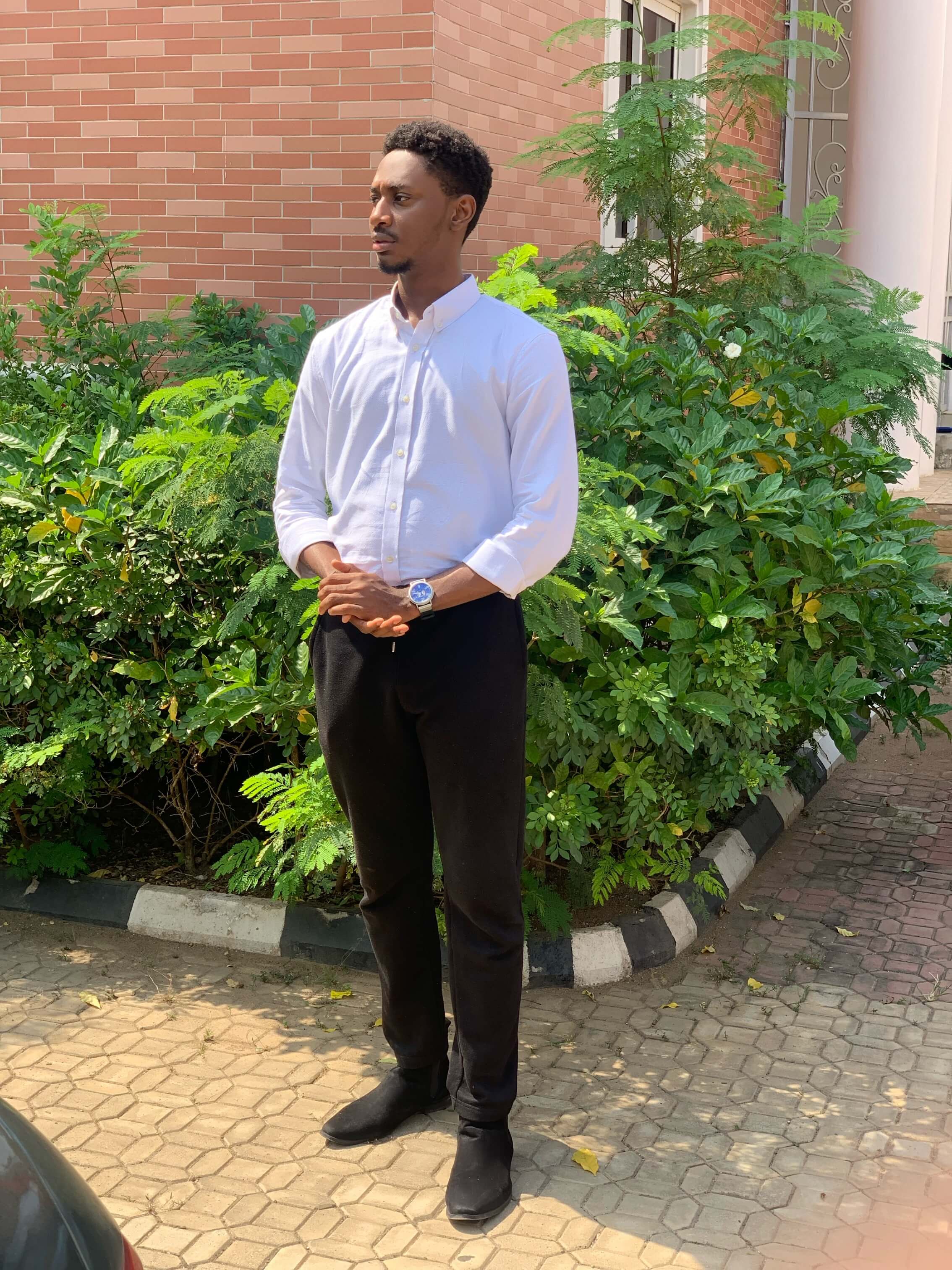 Chinwike Maduabuchi in a button-down white tee, shirt arms cuffed, hands held together hanging above waist, staring off to the side and standing. Green plants behind him.